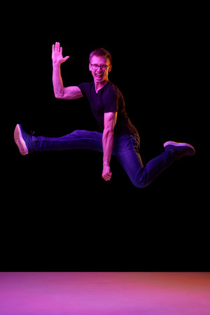 Portrait of young man in glasses posing, cheerfully jumping isolated over black background in neon light. Concept of emotions, facial expression, lifestyle, youth. Copy space for adの写真素材