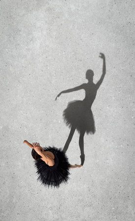 Portrait of young ballerina in black dress performing isolated over grey wall background. Top view. Shadow element. Concept of classic ballet, inspiration, beauty, dance, creativityの写真素材