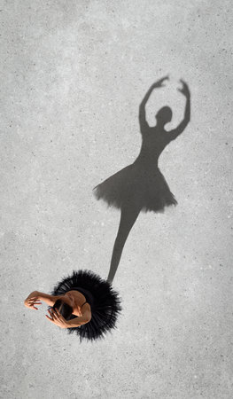 Portrait of young ballerina in black dress performing, standing on pointe isolated on grey wall background. Top view. Shadow element. Concept of classic ballet, inspiration, beauty, dance, creativityの写真素材