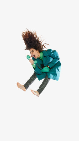 Portrait of young woman in green trench coat jumping on white background, shouting in megaphone in...