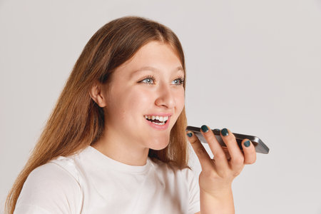 Close-up portrait of young cheerful girl recording voice message on phone isolated over grey...