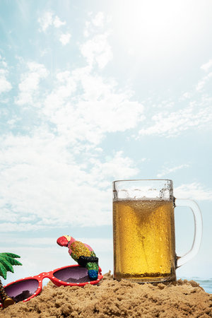 Image of light foamy beer, glass standing into sand over sea and sky background. Summertime chill....