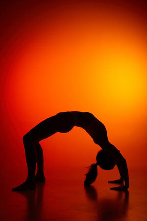 Silhouette of female full-length body isolated over orange background. Standing on bridge yoga pose. Body art, aesthetics. Concept of beauty, femininity, health, sport, lifestyleの写真素材