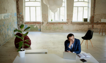 Portrait of young businessman sitting with thoughtful face at the table with laptop in empty room. Failure of project, professional crisis. Concept of business, challenges, work, occupationの写真素材