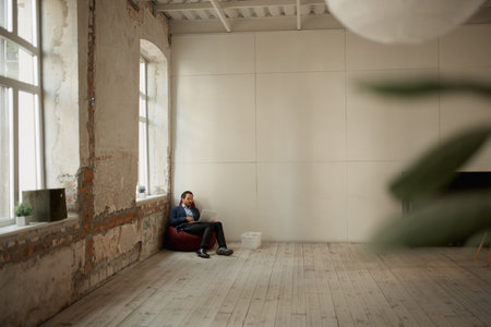 One businessman sitting in the corner of big room on floor pillow with laptop and working. Buying...