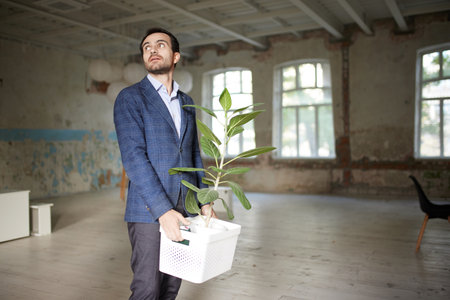 Portrait of young businessman walking out empty room with flower. Business closure. Depression and...