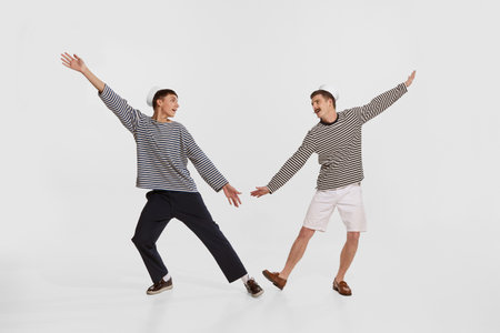 Portrait of two young boys, sailor, seamen in striped shirts dancing on deck isolated over white...