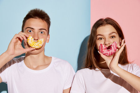 Portrait of young people, man and woman posing with donuts near mouth isolated over blue-pink...