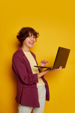 Portrait of cheerful teen girl, student with curly brown short hair posing with laptop isolated over yellow background. Concept of youth culture, emotions, facial expression, fashionの写真素材