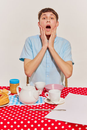 Portrait of young emotive boy in blue shirt sitting at the table and having breakfast isolated on grey background. Concept of emotions, retro style, fashion, 60s, 70s, family. Copy space for adの写真素材