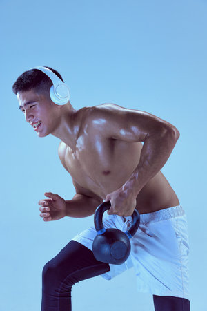 Portrait of young muscular man training in headphones, lifting weight isolated over blue background...