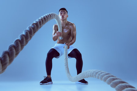 Portrait of young muscular man training, pulling rope isolated over blue background in neon light....