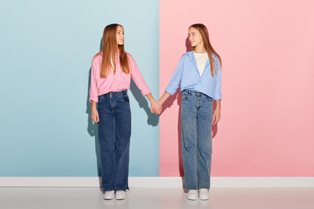 Pink and blue. Stylish couple of teen girls in trendy warm clothes looking at each other isolated over colorful background. Fashion, beauty, emotions, friendship and ad. Look happy, delightedの写真素材