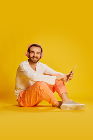Mature handsome happy man with beard in casual clothes sitting on floor isolated over yellow...