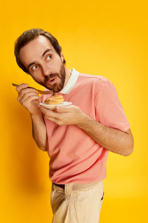 Handsome stylish man with moustache in polo-shirt tasting eclair isolated over yellow background. Facial expression, healthy eating, humor, positive emotions conceptの写真素材