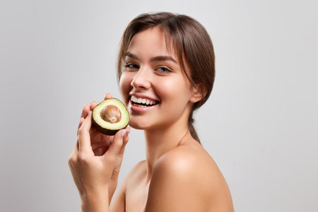 Half-length portrait of happy woman with bared shoulders holding avocado and looking at camera with...