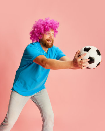 Emotional football fan, young caucasian man in funny wig watching soccer match and expressing emotions over pink background. Sport, fans, footballの写真素材