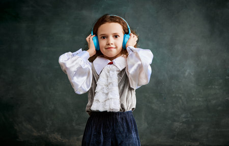 Music. Portrait of little charming girl with curly hair in headphones over dark background. Concept of fashion, comparison of eras, retro, vintage, emotions. Looks happy, delightedの写真素材