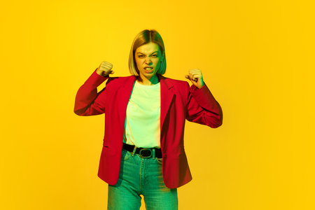 Anger. Annoyed young girl with blonde hair wearing red jacket with vivid facial expression over yellow background. Concept of human emotions, mood, fashion, ad and mental healthの写真素材