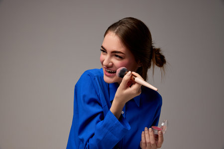 Half-length portrait of young happy smiling beautiful girl holding blush brush and doing makeup...