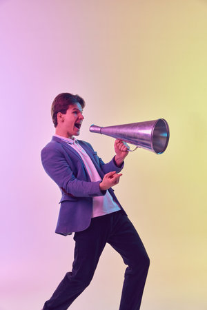 Young excited man shouting in megaphone, having fun isolated over light background in neon light....