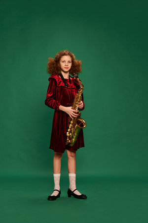 Curly redhaired happy school age girl wearing festive dress playing on saxophone over green background. Studying, fashion, art, music and creative retro style. Looks cute, funnyの写真素材