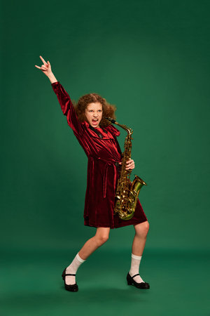 Curly redhaired happy school age girl wearing festive dress playing on saxophone over green background. Studying, fashion, art, music and creative retro style. Looks cute, funnyの写真素材