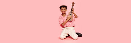 Happy emotional little african boy in retro style clothes and cap posing with ukulele guitar...