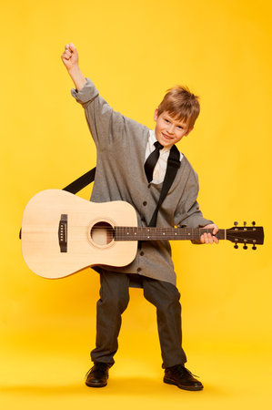 Charming little boy, funny musician wearing huge oversize clothes playing guitar, having fun isolated over yellow background. Pop art, music, new vision, fun concept. Aspiration and creativityの写真素材
