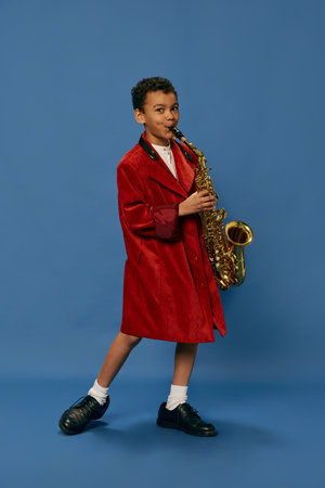 Like jazz man. Portrait of cute little african boy wearing huge mans jacket and shoes playing on saxophone over blue background. Fashion, art, music and creative style. Looks happy, cheerfulの写真素材