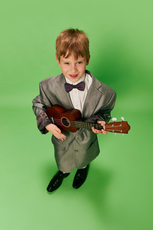 Stylish little school age boy in huge oversize retro style clothes and big male shoes posing with ukulele guitar on green background. Concept of music, childhood, education, fashion. Wide angle viewの写真素材