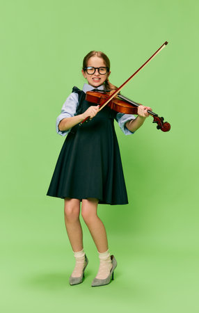 Cute little girl, talanted musician wearing huge mothers heels shoes playing violin, having fun isolated over green background. Pop art, education, music, classic art, new vision, fun conceptの写真素材