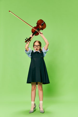 Cute little girl, talanted musician wearing huge mothers heels shoes playing violin, having fun isolated over green background. Pop art, education, music, classic art, new vision, fun conceptの写真素材
