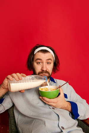Healthy eating. Funny mature bearded man wearing homewear, pijamas has breakfast over bright red background. Positive emotions, home cozy atmosphere, funny meme emotions, adの写真素材