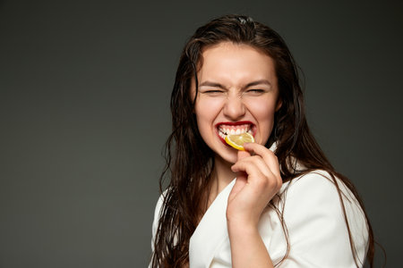 Funny whitening. Emotional fashionable stylish beautiful woman wearing white jacket tasting slice of orange over gray background. Concept of healthy lifestyle, vitamins, dieting, fashion, beautyの写真素材
