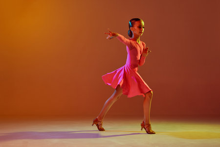 Solo performance. Studio shot of little girl, 11 years old kid in festive stage dress dancing ballroom dance over brown background in neon light. Concept of beauty, professional dancesの写真素材