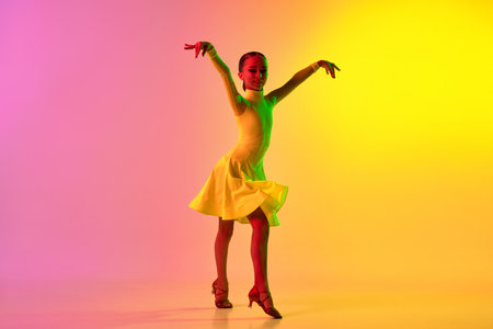Music in movements. Emotional little girl in yellow stage dress dancing classical ballroom dance over gradient pink-yellow background in neon light filter. Concept of beauty, professional dancesの写真素材