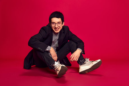 Cheerful, joyful. Stylish handsome man with glasses posing sitting on floor and smiling over red background. Concept of education, studying and student lifeの写真素材