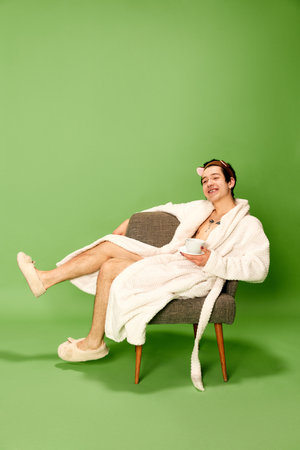 Relax time. Portrait of relaxing young man wearing white robe and sitting on chair with cup of coffee over green background. Concept of positive emotions, happinessの写真素材