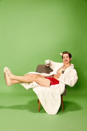 Young man wearing female bathrobe and slippers sitting on chair with cup of tea over green background. Concept of vacation, self love and self care, weekend, rest, relax. Weird people cの写真素材