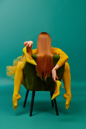 Creative portrait of redhead girl with long straight hair posing on armchair, sitting in strange poses over cyan color background. Impersonal emotions, body language. Fashion, beauty, mental healthの写真素材