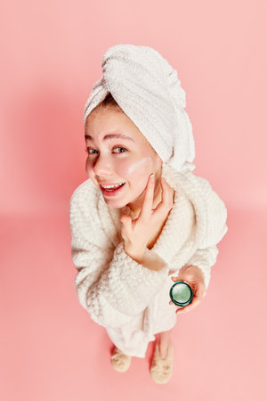 Morning beauty routine. Top view of cute beautiful girl wearing bathrobe shoulders using face cream to moisturize skin over pink background. Concept of beauty, natural make-up, skin care, healthy lookの写真素材
