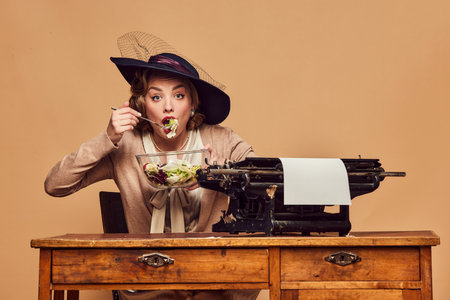 Busy and hungry. Charming woman wearing hat sitting at typewriter and eating salad over beige background. Concept emotions, beauty, fashion, retro style, vintage, 60s, poems, novelの写真素材