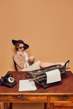 Lunch break. Adorable woman writer wearing heels and glasses sitting with legs up on table eating snacks over beige background. Concept emotions, beauty, fashion, retro style, vintage, 60sの写真素材