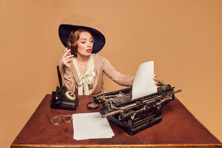 Thoughts, ideas. Beautiful woman writer sitting at typewriter smoking and reading poems over beige background. Concept poems, novel, emotions, beauty, fashion, retro style, vintageの写真素材