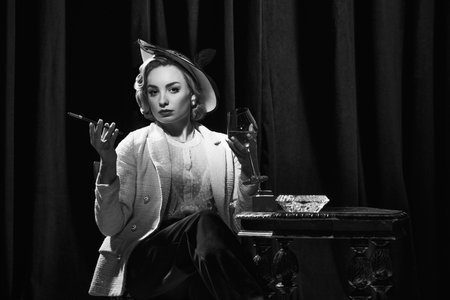 Calm, pensive. Charming woman wearing retro clothes sitting at table holding wineglass and smoking. Concept of beauty, fashion, old films, actress, retro, vintageの写真素材
