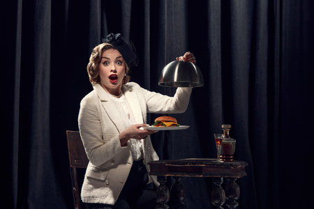 Time to eat. Charming woman wearing vintage costume holding burger with surprised face and open mouth. Concept of human emotions, beauty, fashion, old films, actress, retro, vintage, foodの写真素材