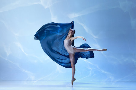 Showing flexibility and grace. Ballerina wearing beige bodysuit dancing elegant movements with silk over blue background. Concept of classic ballet, inspiration, beauty, dance, creativityの写真素材