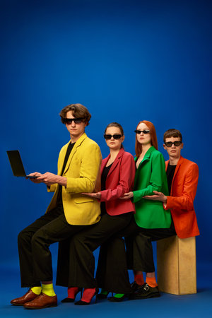 Common support. Young team wearing colorful jackets sitting on each other knees and working over blue background. Concept of fashion, models, style, business, online, teleworking, promotionの写真素材