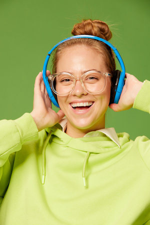 Blonde girl smiles. Closeup happy young girl wearing glasses and wireless headphones listening to music and smiling over green background. Concept of emotions, happiness, fashion, youth, ad and musicの写真素材